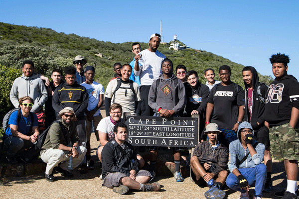 A group of young men stand around a latitude and longitude sign in Capetown South Africa.