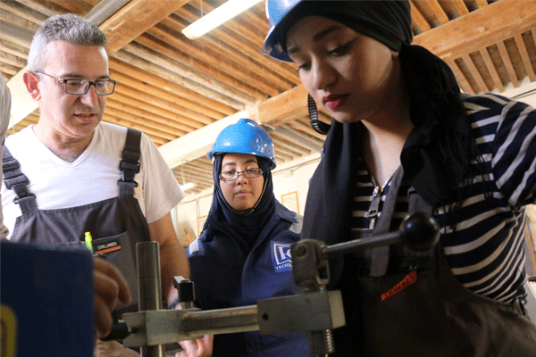 A woman works at a small machine while a man and another woman look on behind her.