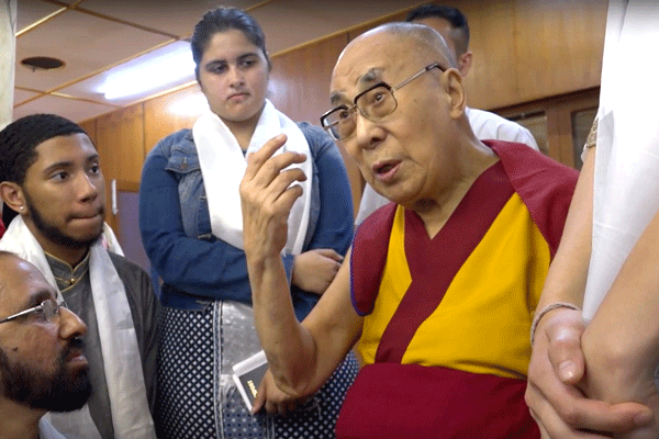 The Dalai Lama sits and speaks to several young people gathered around him.