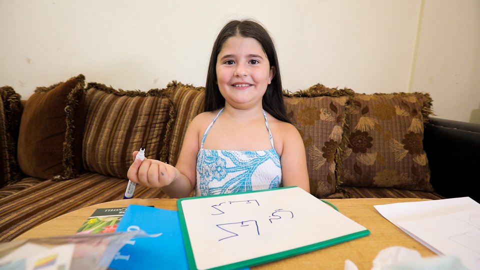 A young girl holds up her marker after writing in Arabic on a sheet of paper on the table in front of her.