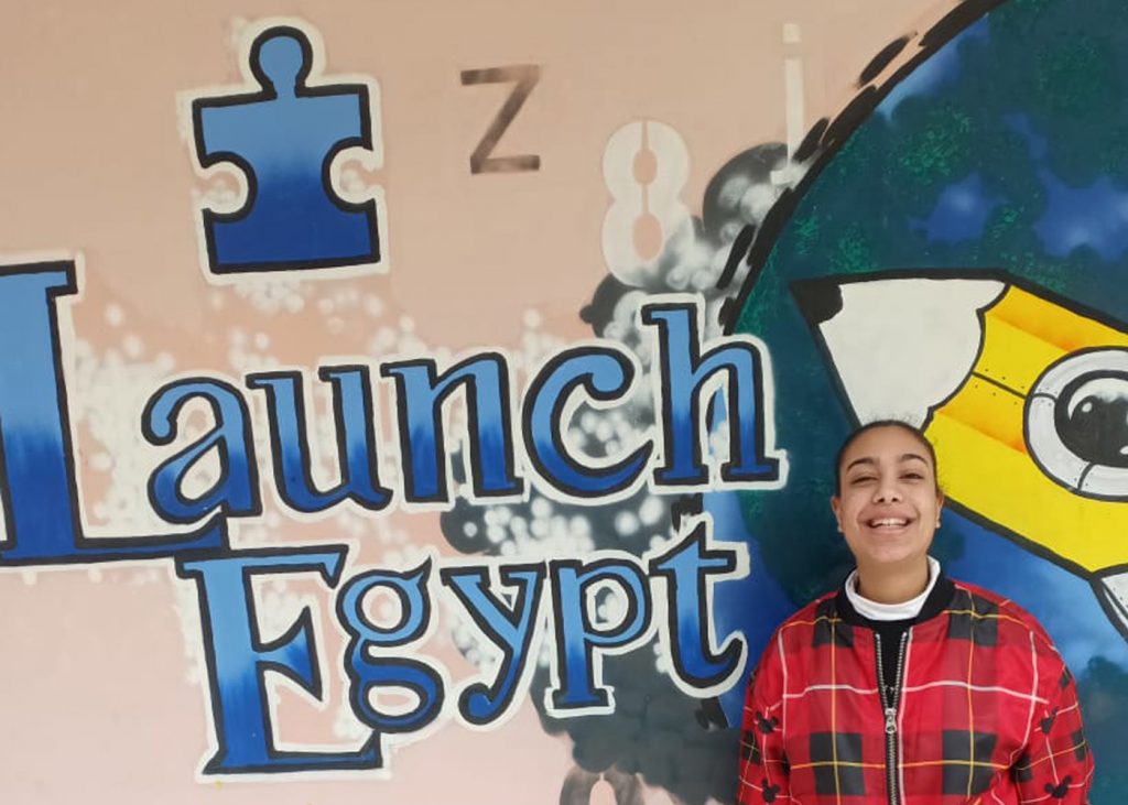 A student smiles in front of a sign that reads 'Launch Egypt' in blue on the Launch Egypt campus in Cairo, Egypt