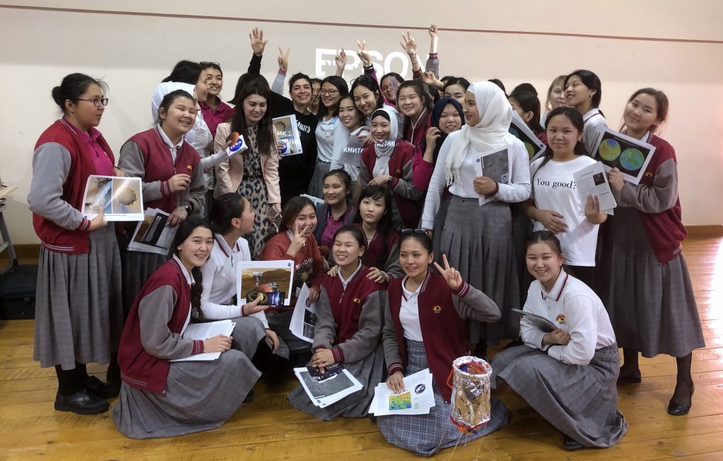 A group of girls in uniform and one adult pose while holding up photos and certificates.