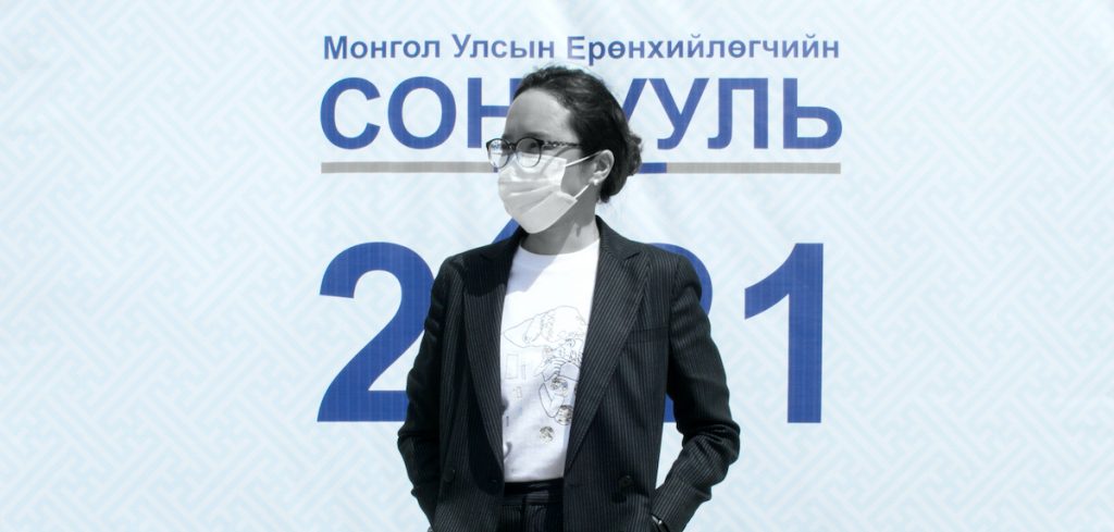A woman stands in front of a sign for the Mongolian 2021 election