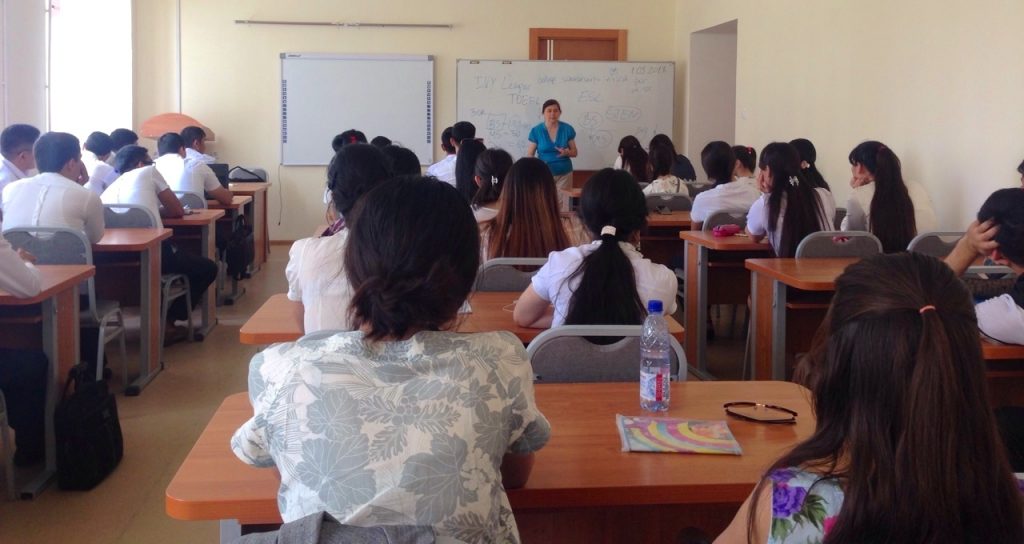 A woman speaks in front of a white board in front of a classroom full of students.