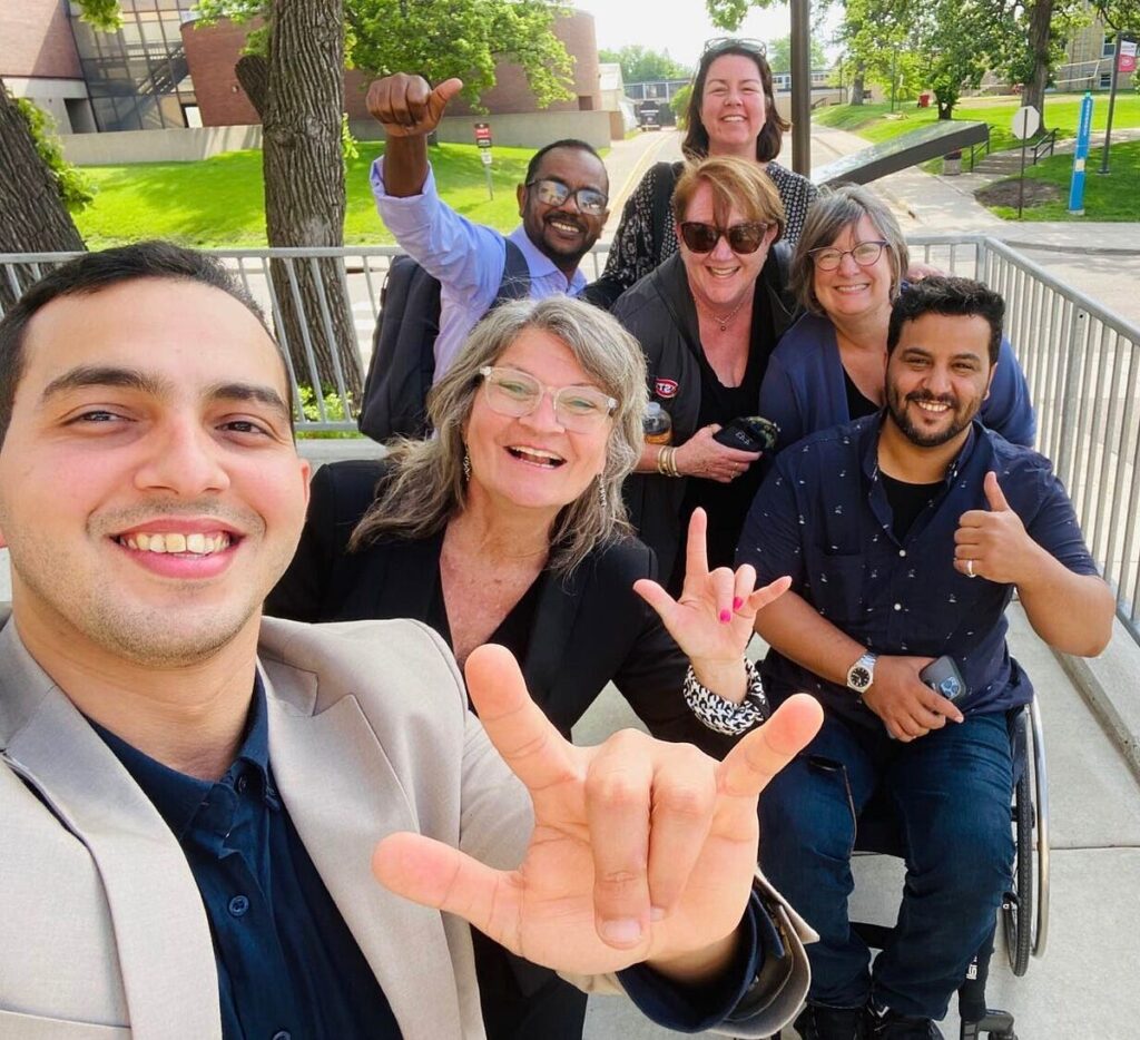 A group selfie of seven individuals. One of them is in a wheelchair. Two of them are holding up the American Sign Language symbol for "I love you" and two others are giving thumbs up.
