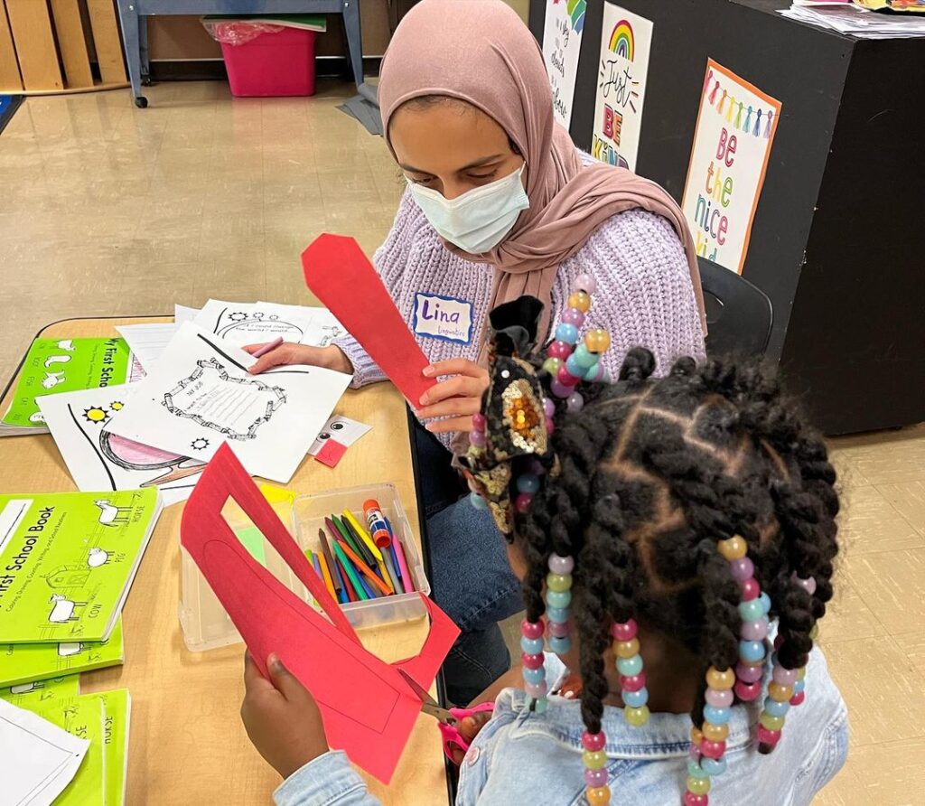 A person wearing a pink hijab, pink sweater, and white face mask sits at a desk covered in arts and crafts supplies. The person is holding a red piece of paper for a child who is next to her, wearing colorful braids.