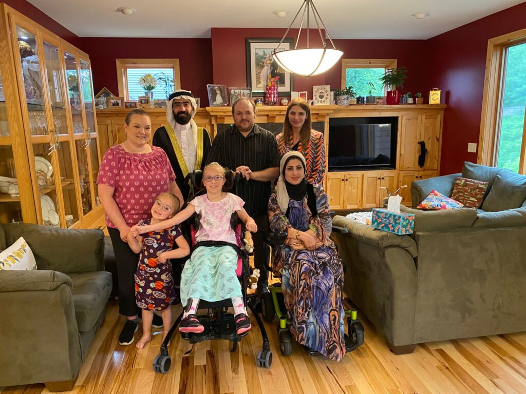 Five adults and two children are in a living room together. Five are standing while two are in a wheelchair.