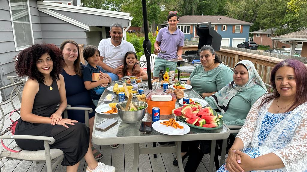 Eight individuals are sitting and one is standing at an outdoor patio table. On the patio table is a range of food items such as watermelon, corn cobs, and salad.