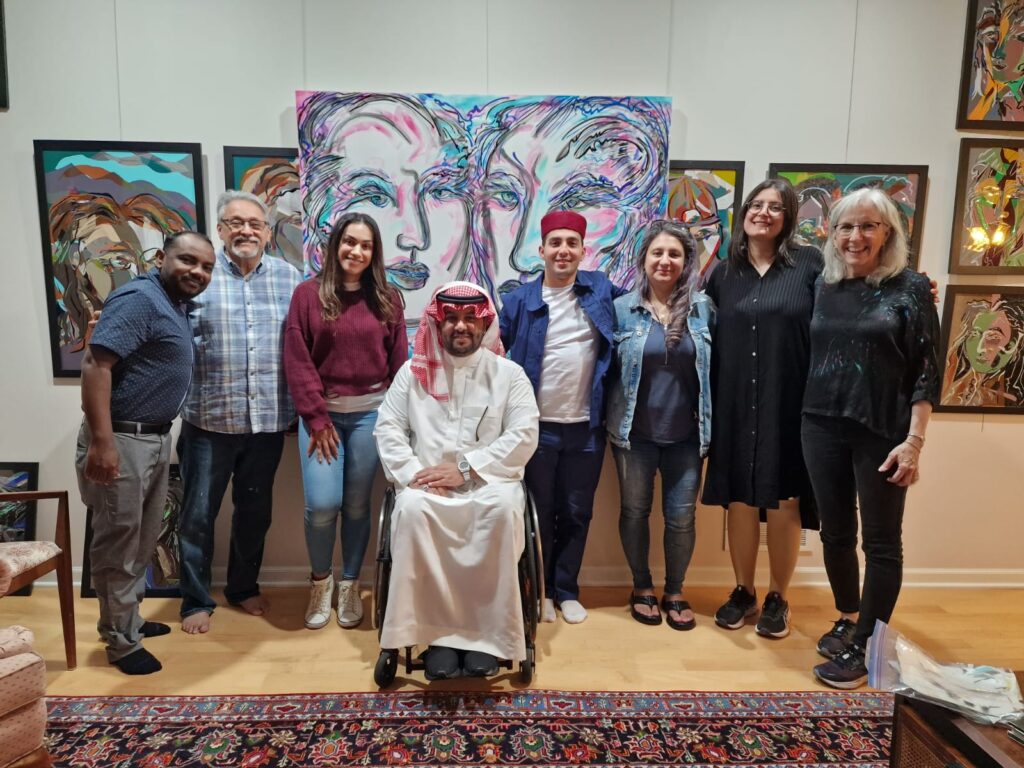 Eight individuals together in an art gallery with paintings on the wall lined up behind them. One of the individuals is in a wheelchair.