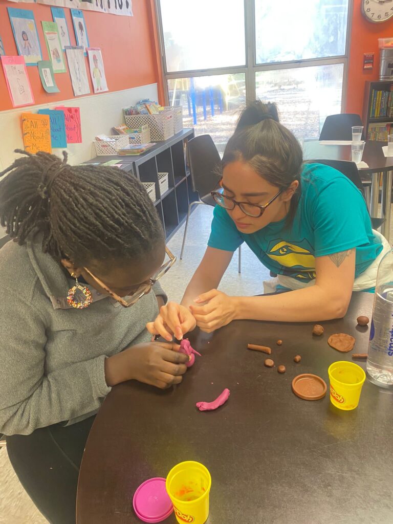 Two individuals work together at a table doing arts and crafts involving play-doh. The person on the left is wearing a gray sweater and colorful glasses and earrings. The person on the right is wearing a short-sleeved teal shirt and black glasses.