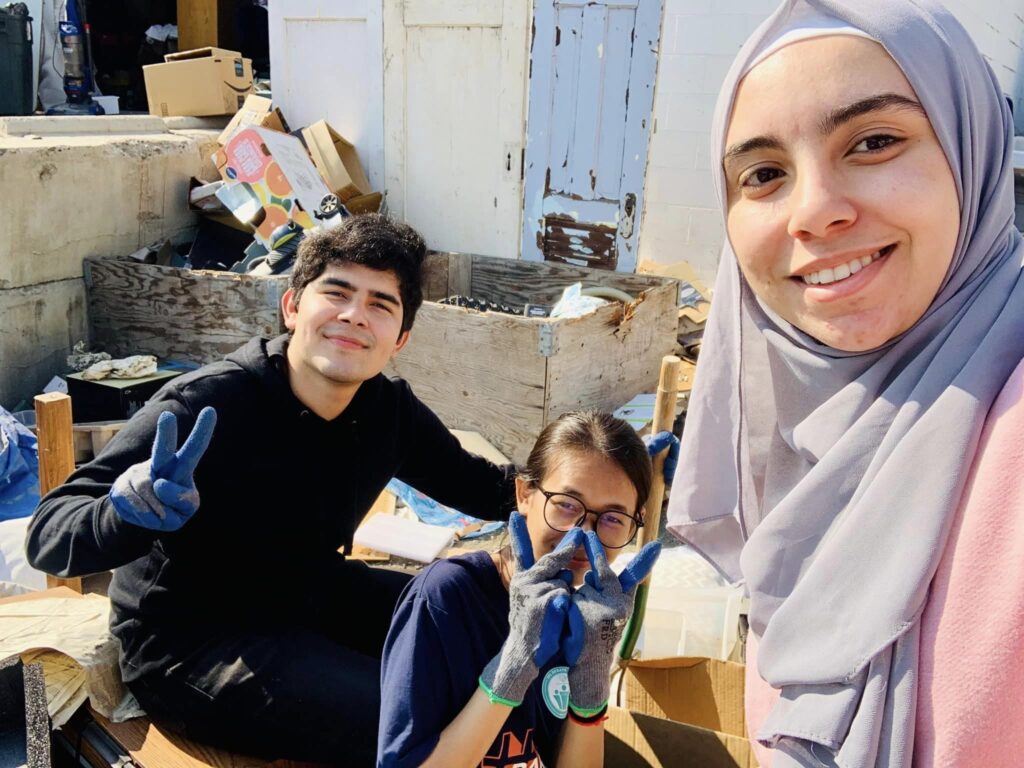 Three people pose together at an outdoor community service location. The person on the left is wearing a black sweater and blue work gloves. The person in the middle is wearing a blue short-sleeved shirt, black glasses and blue work gloves. The person on the left is wearing a pink top and a light gray hijab.