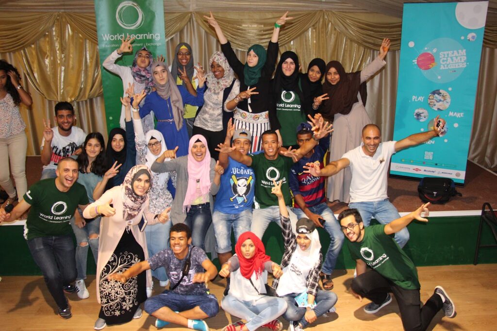 Group of 23 students in 3 rows posing in from of a tall World Learning banner and a STEM Camp Algiers banner. They are smiling and making funny faces and have their arms outstretched, making peace and ‘okay’ signs with their hands. They are approximately high school age.