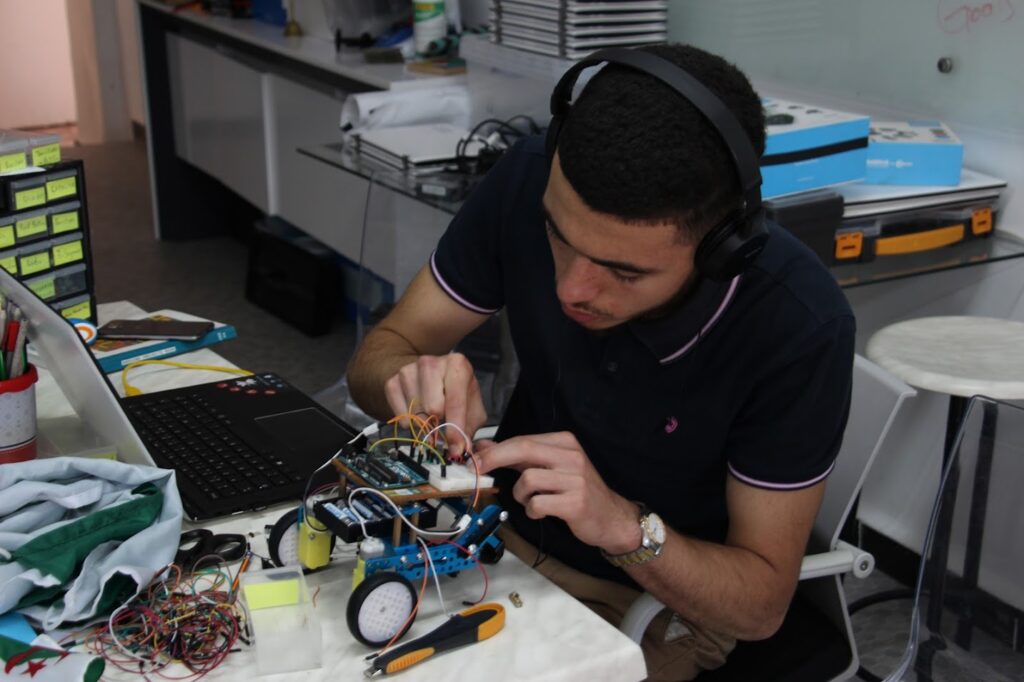 A male student sitting in front of a laptop and fixing a robot on the table. He is approximately high school age.