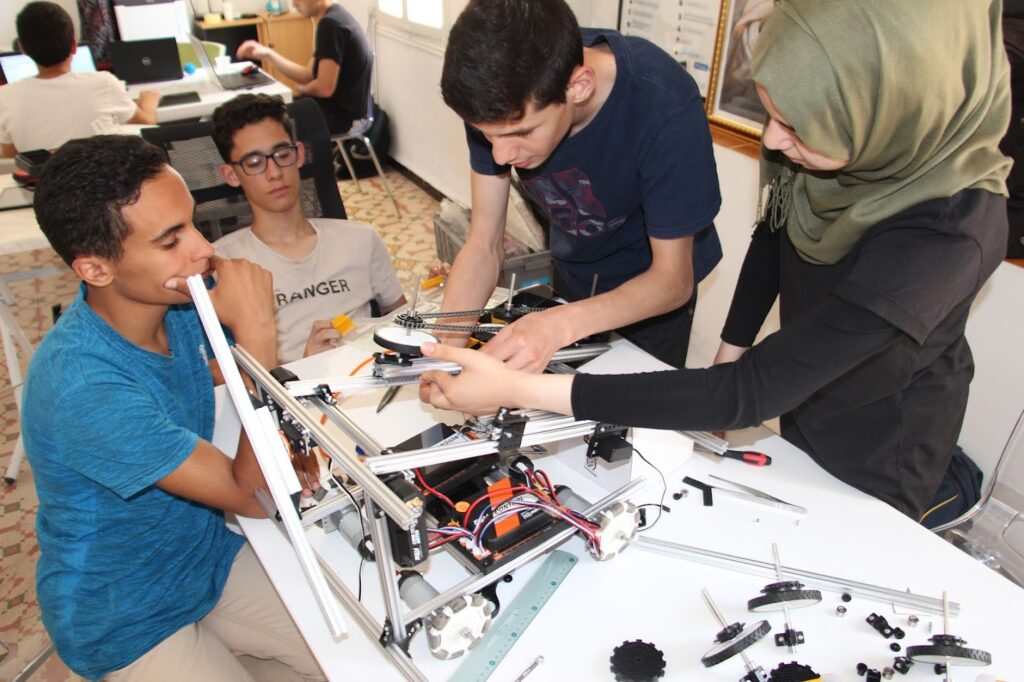 Four students, three male and one female, working on a large robot device that cover the end of a table. They are approximately high school age.