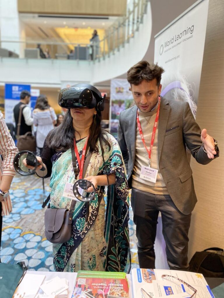 A female is wearing a virtual reality helmet over her eyes and holding two virtual reality handsets. She is standing next to a male who is explaining the device to her.