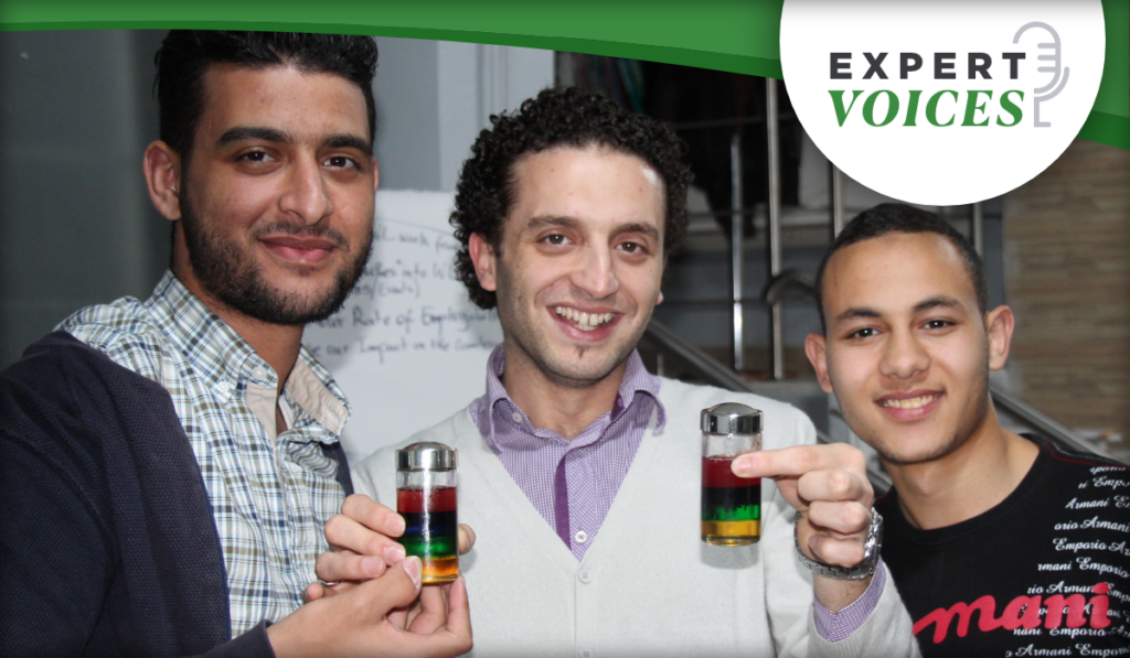 Three males facing the camera. Two are holding laboratory vials that have red, green, and yellow liquid.