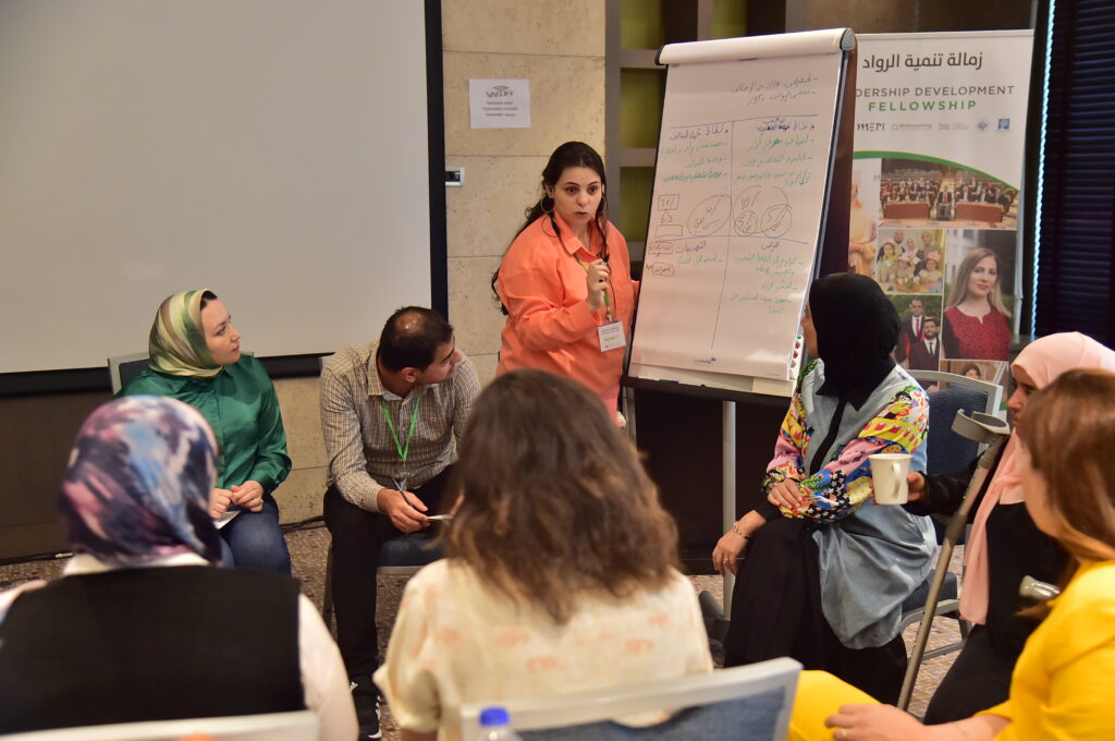 Seven fellows sit and one stands around a large white notepad and discuss their negotiation strategies during a simulated negotiation activity.