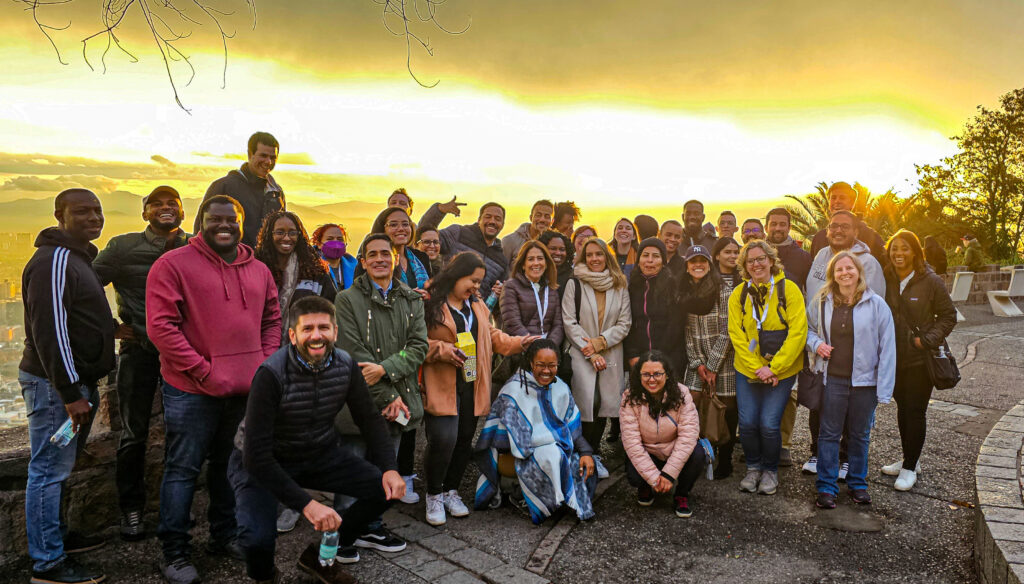 A group of 35 people posing in a huddle at the top of a hill. The sky is yellow behind them, and they are wearing jackets and coats.