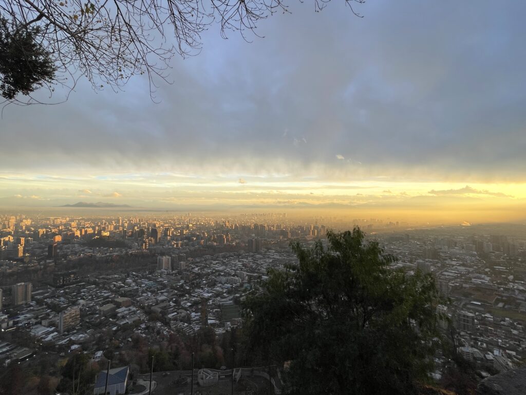 View of a sunset from the top of a hill looking down on a large city. The sky is cloudy and the horizon is yellow.