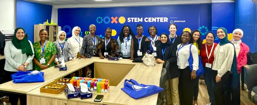 Sixteen women are standing in a semicircle around a large wooden table. They are smiling and standing before a blue sign that says “STEM Center.”