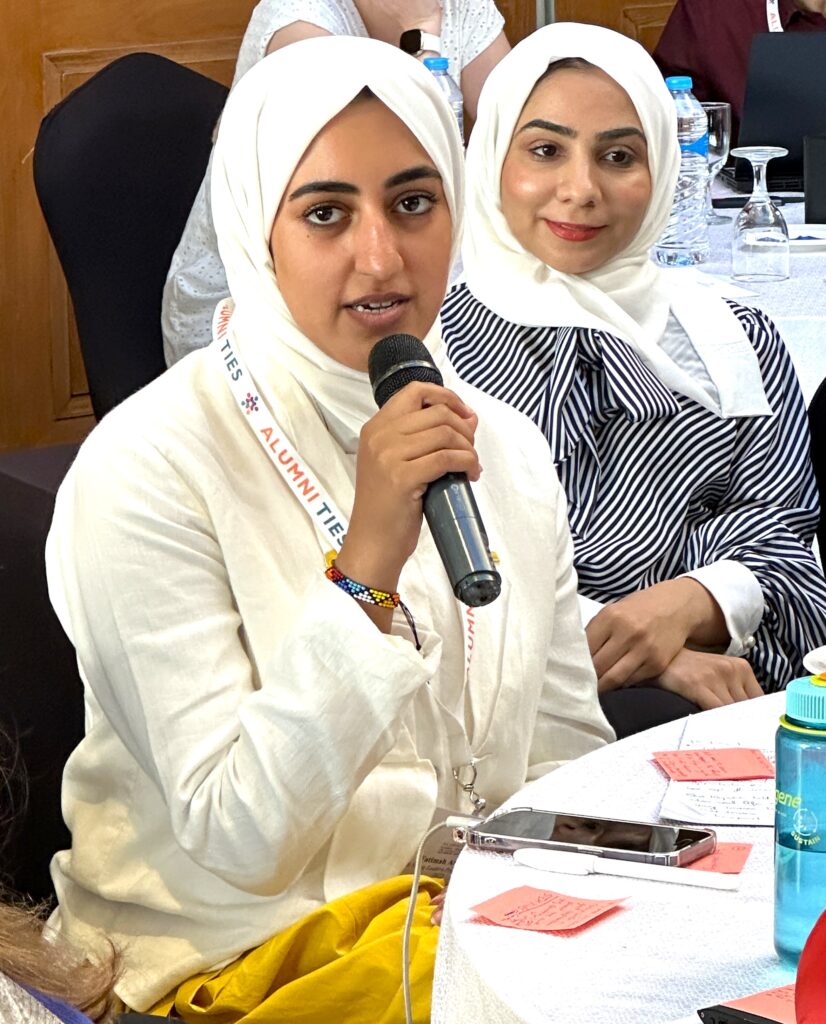 A woman wearing a white hijab is sitting at a table and speaking into a microphone. Another woman in a white hijab is sitting behind and listening to her.