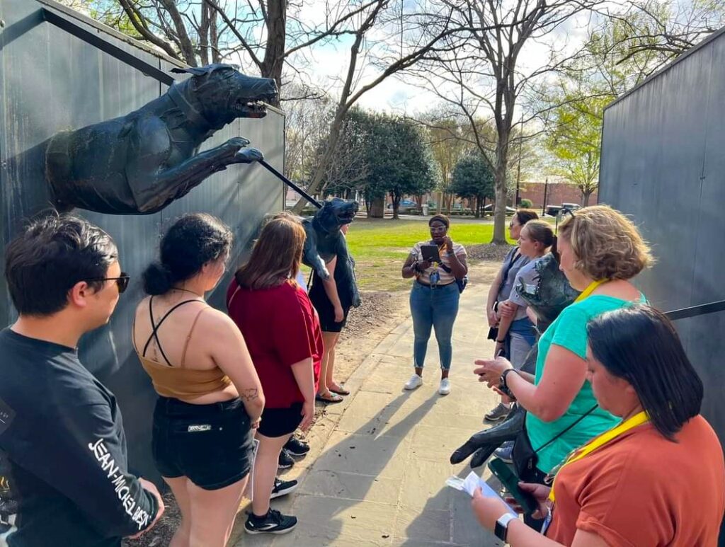 A group of nine students are standing between the walls of a black metal monument dedicated to those who fought for civil rights. The monument has two metal dogs leaping off the wall.