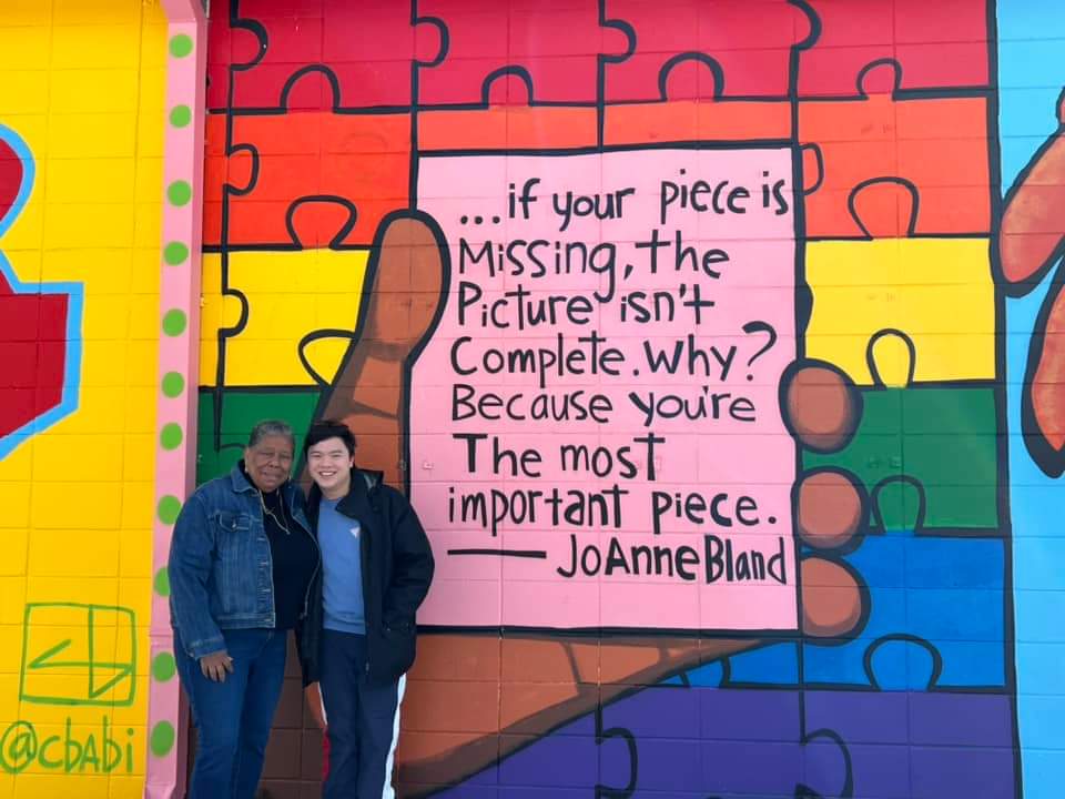 An older Black woman standing next to college student from Singapore in front of a mural on a brick wall. The mural is in bright colors and shows a large hand holding a card with a inspirational quote by the woman.