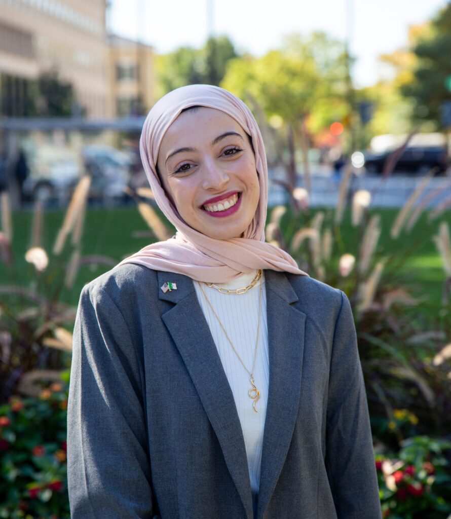A woman in her 20s wearing a pink hijab is smiling at the camera, shown shoulders up. Behind her is a city park and buildings.