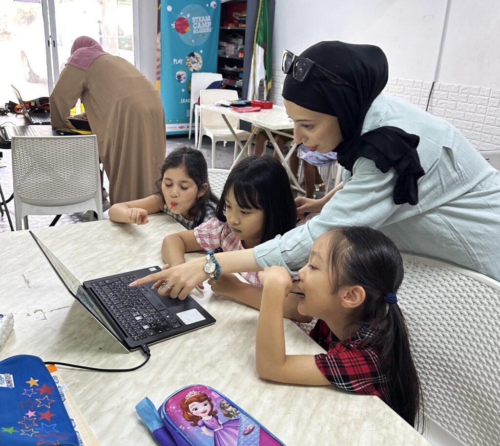 Three elementary school girls are sitting around a laptop. A woman is leaning over them and pointing at the laptop as she provides instruction.