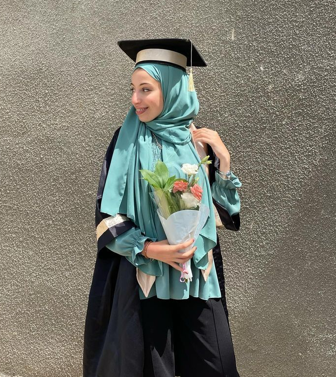 A woman in her 20s wearing a blue hijab and a graduation cap and gown is posing in front of a cement wall. She is looking off to the side, smiling, and holding a bouquet of flowers.