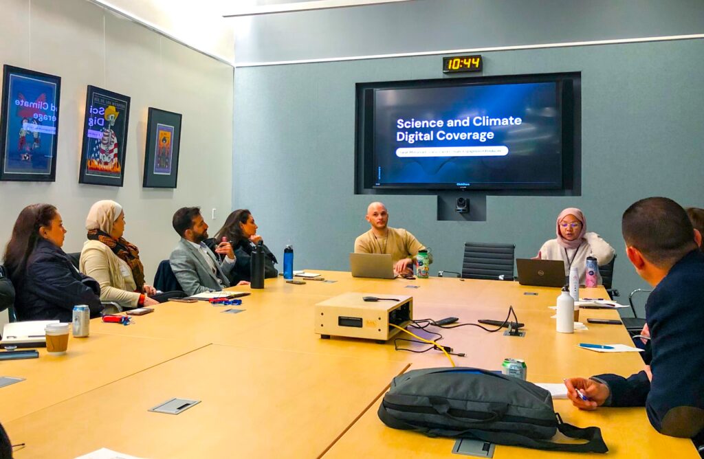 Adults sitting at a long rectangular conference table listening to a presentation on a wall monitor titled "Science and Climate Digital Coverage."