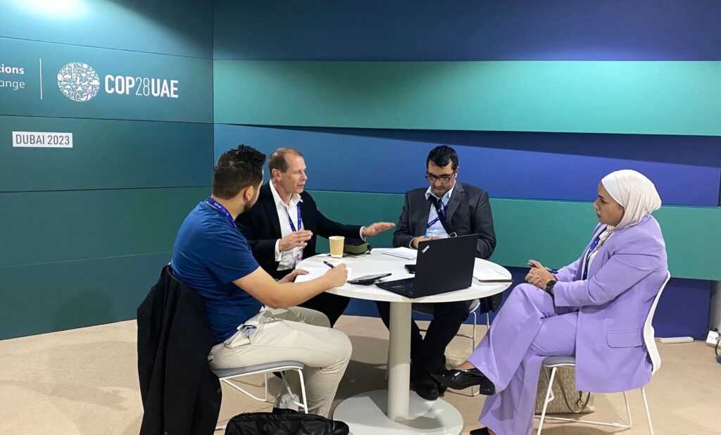 Four men and one woman sitting around a circular table talking. The COP 28 logo is on the wall behind them.