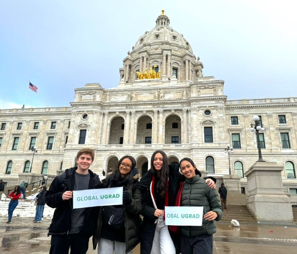 Four Global UGRAD students pose, holding cardboard signs that say Global UGRAD. Behind them is a large white stone building with a dome. Snow is on the ground.
