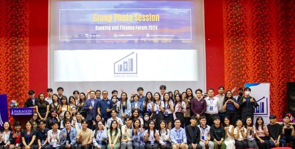 A Global UGRAD alumna poses with approximately 50 fellow students on a stage. A large projector screen is behind them.
