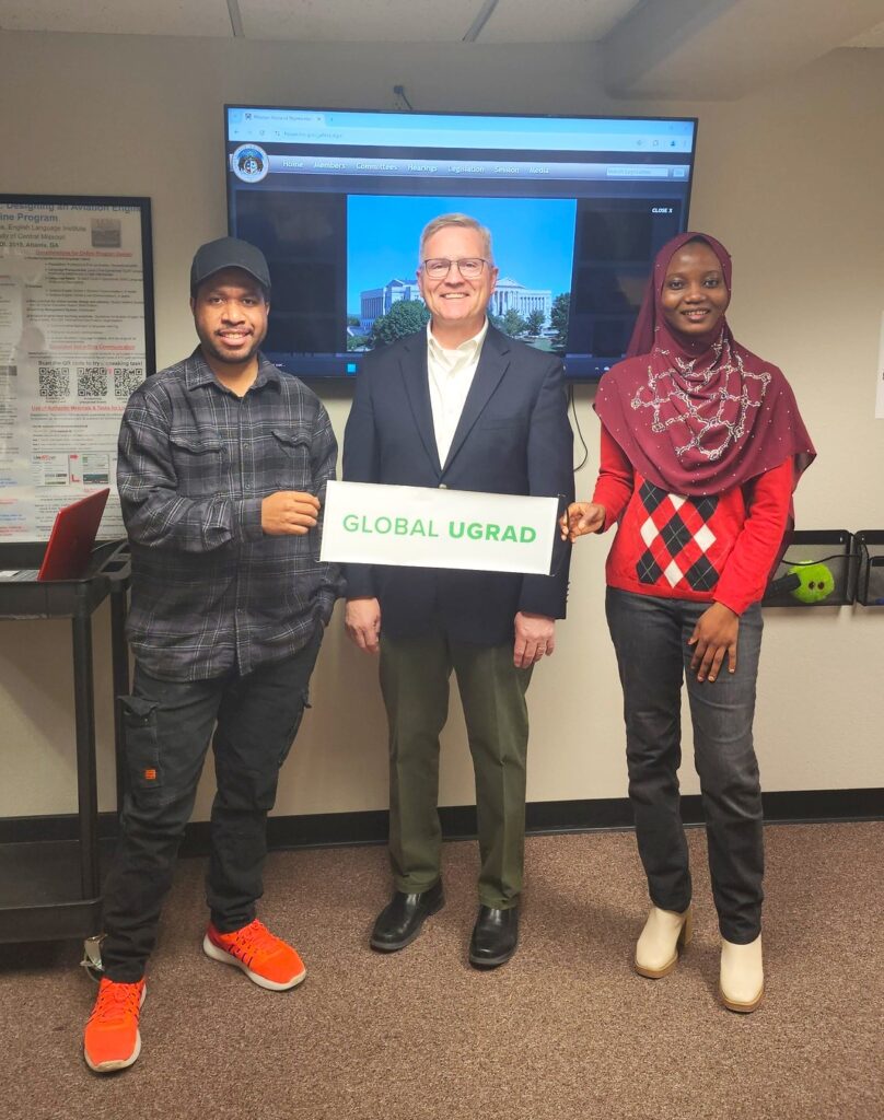 Two Global UGRAD students pose with a man between them. They are holding a cardboard sign that says Global UGRAD. Behind them is a television.