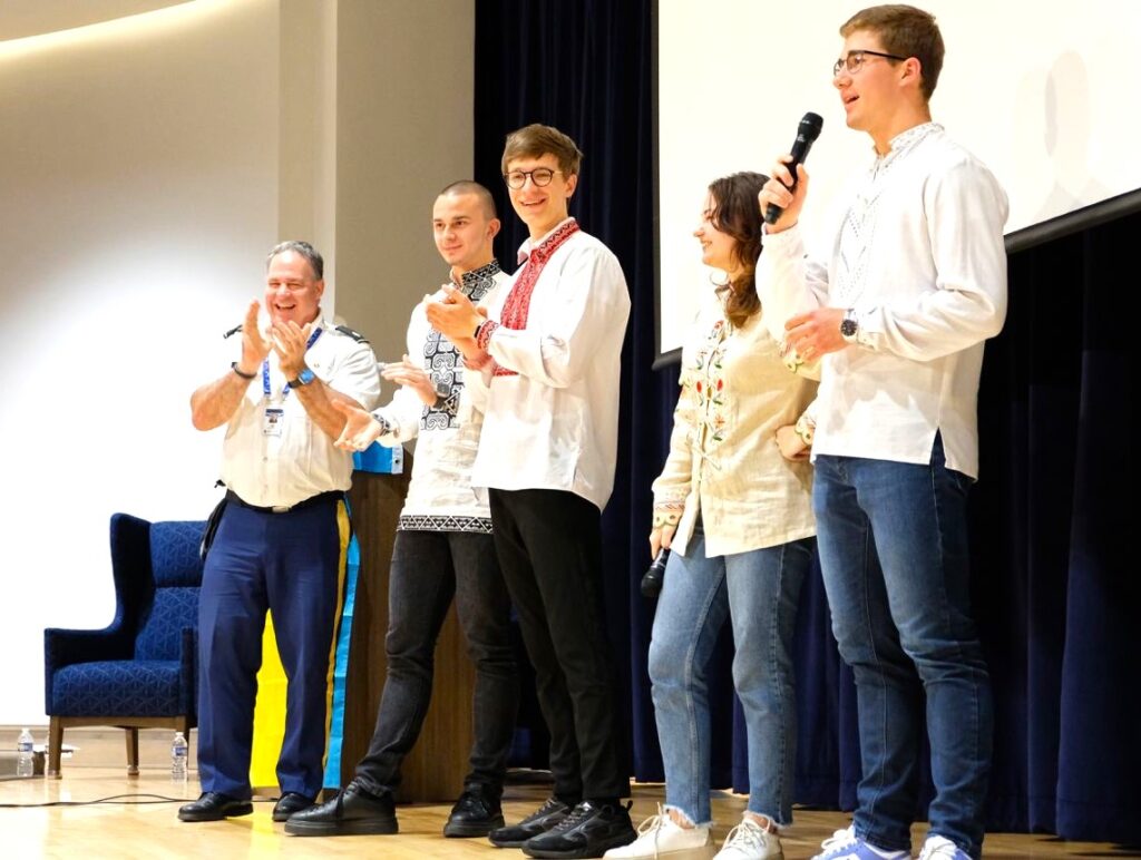 Four Global UGRAD students stand on a stage. One is holding a microphone. A man dressed in a military uniform stands next to them and is clapping.