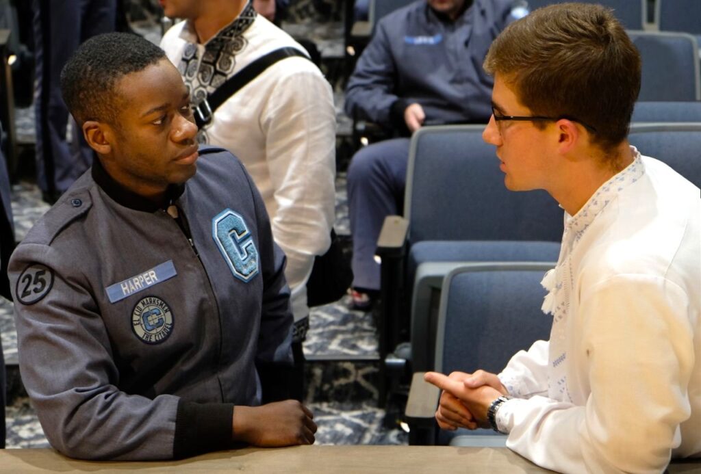 A Citadel cadet and a Global UGRAD student lean on the edge of a stage, talking face to face. One is wearing a military uniform. The other is wearing a white shirt and glasses.