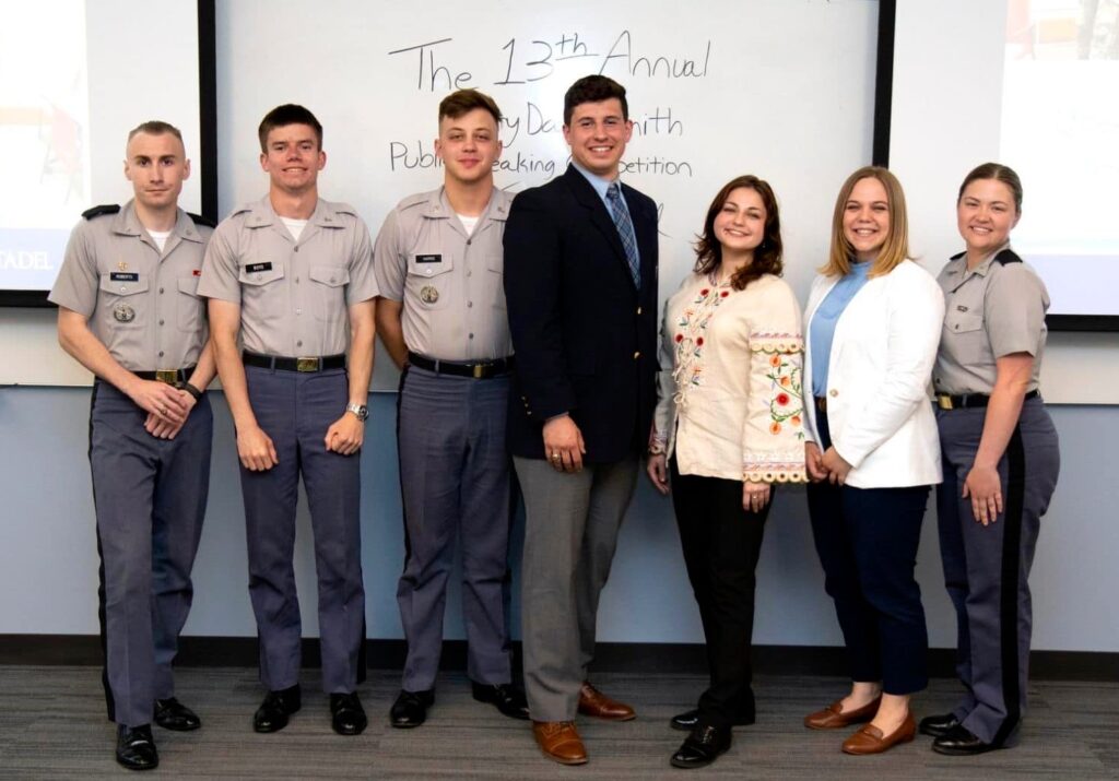 A Global UGRAD student, wearing a traditional top with flowers on it from Ukraine, poses with four Citadel students (one female and four males) all in military uniforms. There are two Citadel faculty members with them wearing business clothing. They all stand in front of a white board.