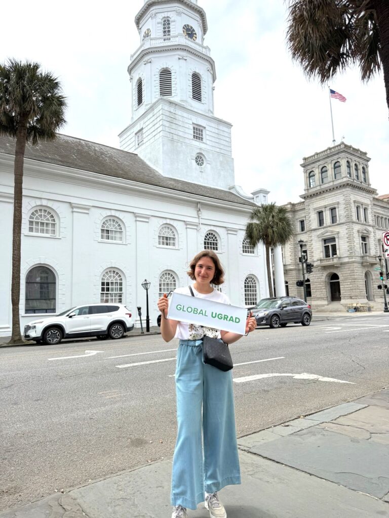A Global UGRAD student stands in front of two white stone buildings with historic architecture. One has a flag at the top. She holds a sign that says Global UGRAD.