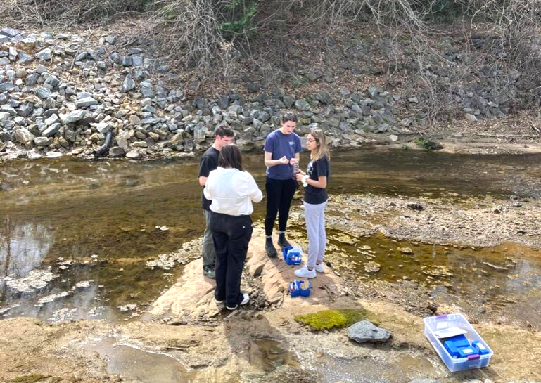 Global UGRAD student stands with three other students at the edge of a creek, holding water sample supplies. A water sample kit is on the ground.