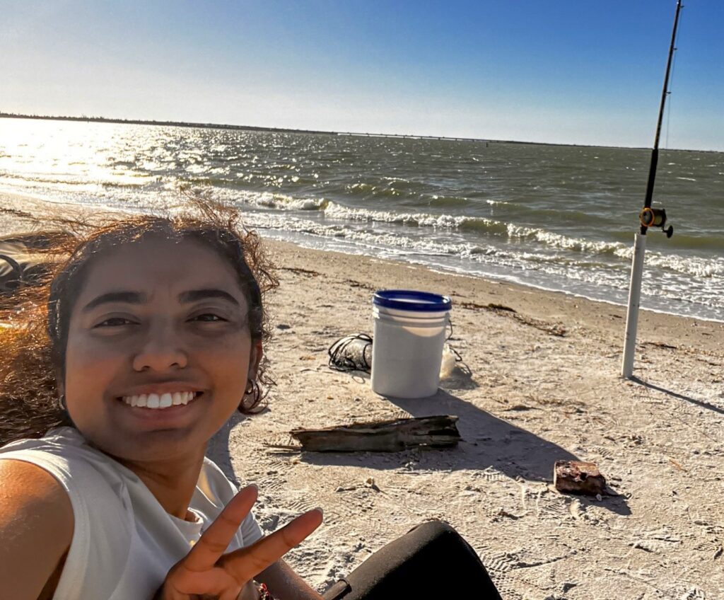 A Global UGRAD student is giving the peace sign with the ocean behind her. On the sand is a bucket, driftwood, and a fishing pole.