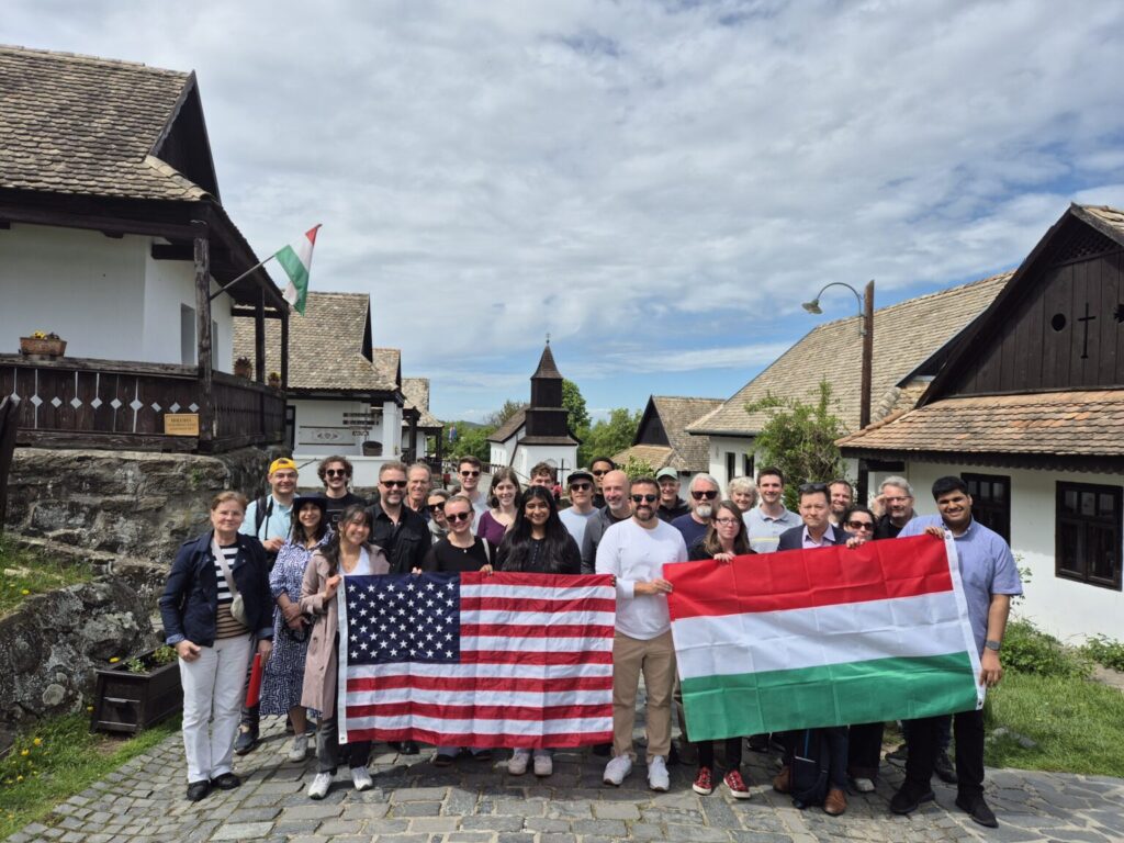 Twenty-five adults stand in a group in front of a series of small village homes and are holding large flags from the U.S. and Hungary.