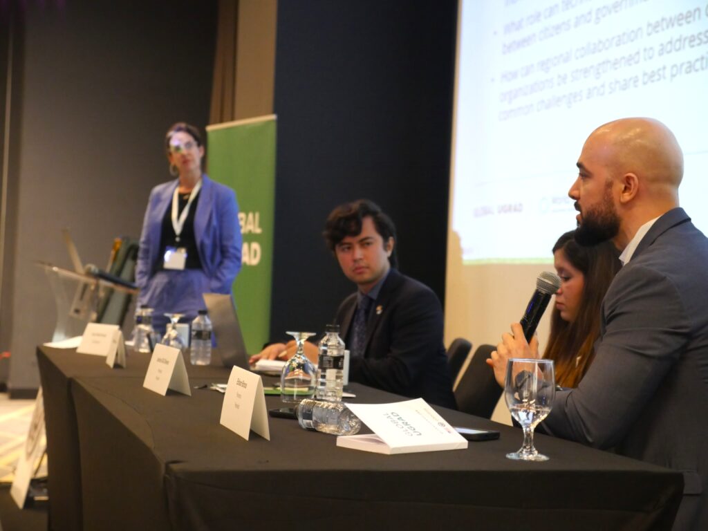 A man, a Global UGRAD alum, is speaking into a microphone while sitting behind a table. Two others are at the table and a women stands at the end of the table. There is a jumbo screen behind them.