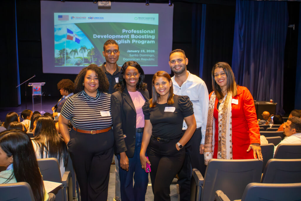 Five World Learning program participants (four women and two men) pose in an auditorium. Behind them, a large screen projects a presentation.