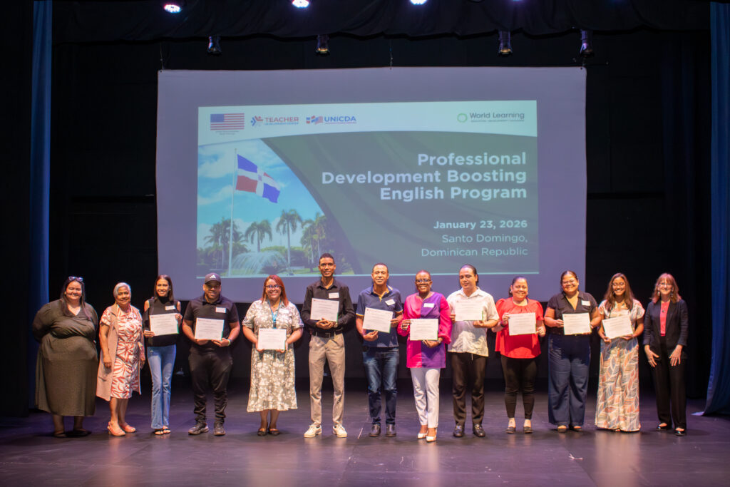 Thirteen adult teachers stand on a stage; ten are holding certificates. A large screen projects a presentation behind them.