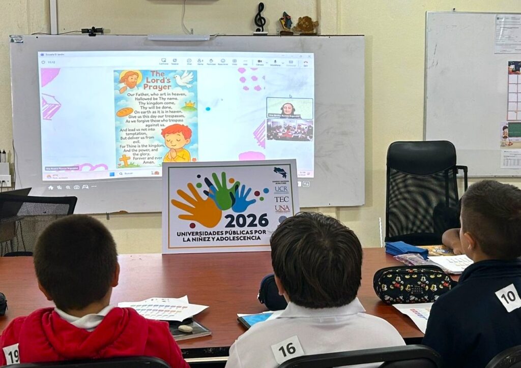 Elementary-aged children sit at a table and look at a presentation projected onto a large screen.