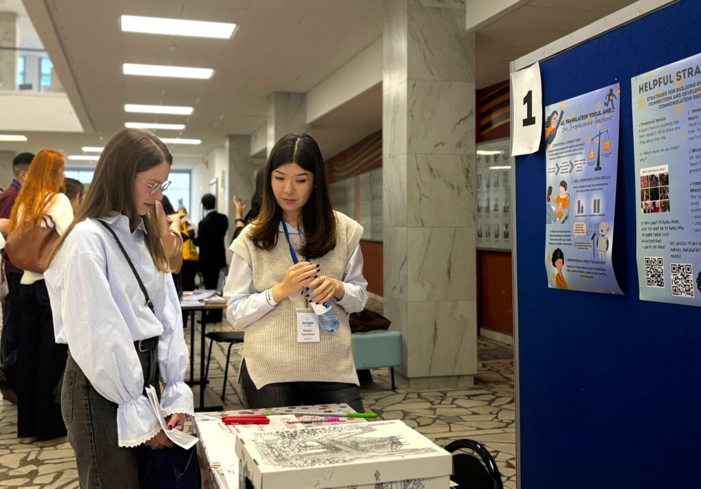A Global UGRAD alumna talks to another female at a small table covered with materials. Two posters hang on a partition near them.