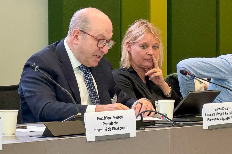 An adult Fulbright Specialist is at a table. A woman is next to him. They have microphones and laptops on the table in front of them, as well as table name tags.