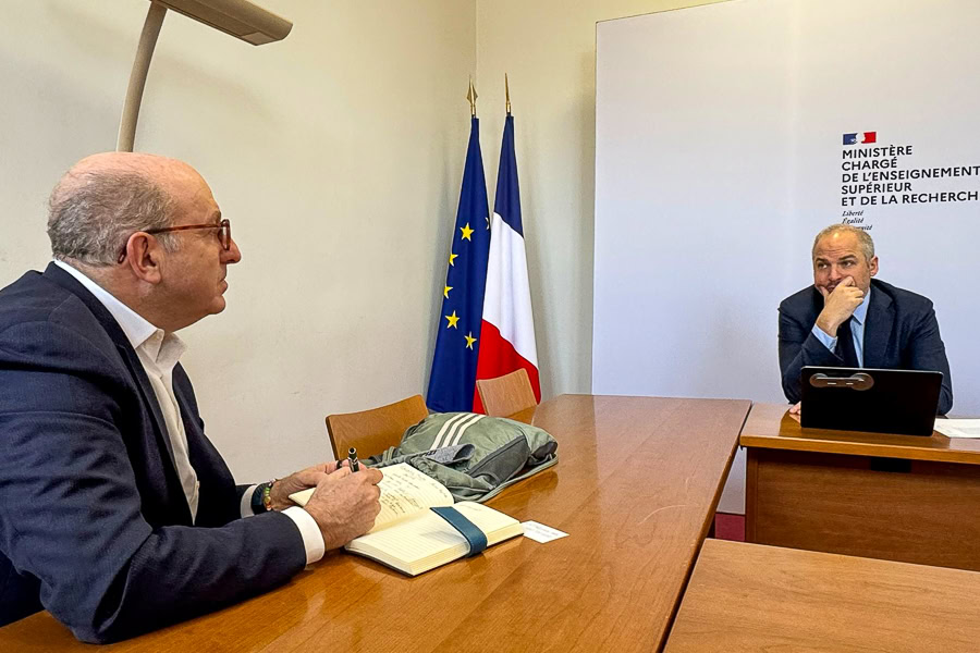 Two men sit at a conference table. One is writing in a notebook while having a conversation. A set of flags is in the corner of the room.