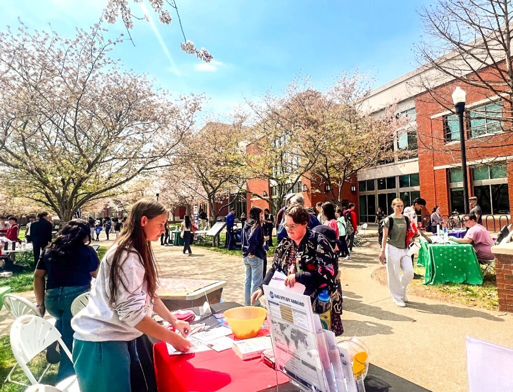 Two women stand at a display table outside on a college campus. Around them are blooming cherry blossom trees.